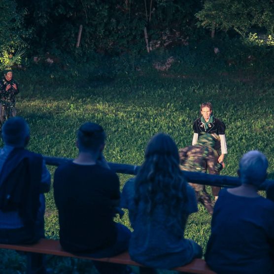 Publikum schaut sich das Theaterstück, das auf einer Wiese stattfindet, auf einer Festbank sitzend