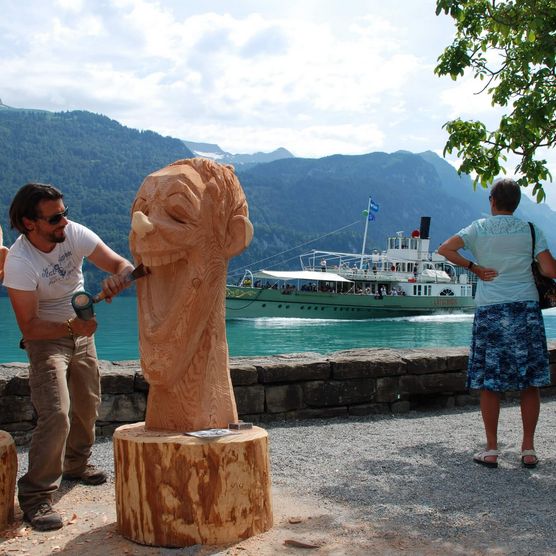 Künstler schnitzt an Holzfiguren vor dem türkisen Brienzersee mit einem Schiff