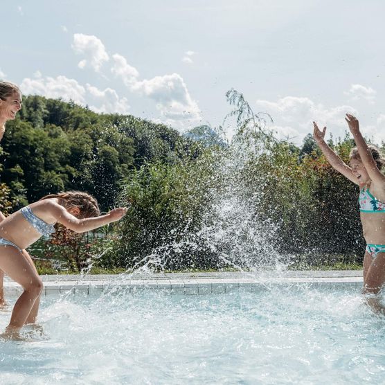 Töchter machen eine Wasserschlacht im Burgseeli während Mutter lachend zuschaut