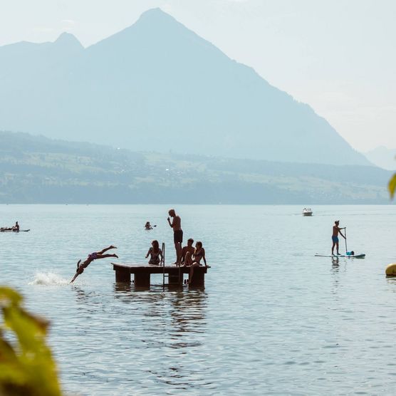 Schülerbad Neuhaus mit Personen, die auf dem Floss stehen oder in den Thunersee springen sowie Stand-up-Paddler und Boote im Hintergrund