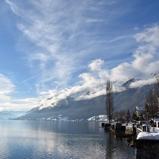 Verschneiter Brienzer Quai entlang des Brienzersees. Im See spiegel sich sich die Wolken, der blaue Himmel und die umliegenden Bergflanken.