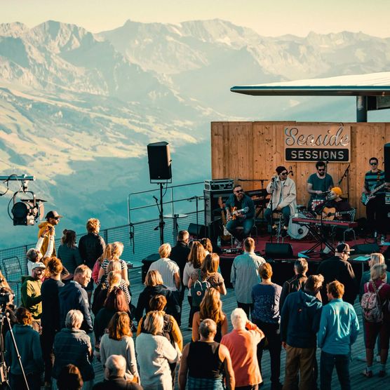 Publikum schaut stehend einem Konzert auf einer kleinen Bühne beim Berghaus Niesen vor einem einzigartigen Bergpanorama