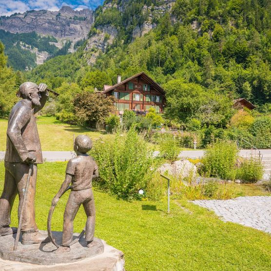 Mitten in der Naturidylle befinden sich handgeschnitzte Holzfiguren auf dem Schwander Lebensweg. Im Hintergrund ein Chalet.